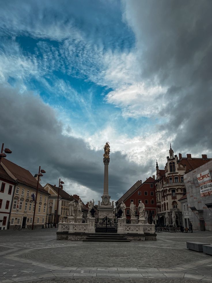 a statue in the middle of a square with buildings around it and clouds above them