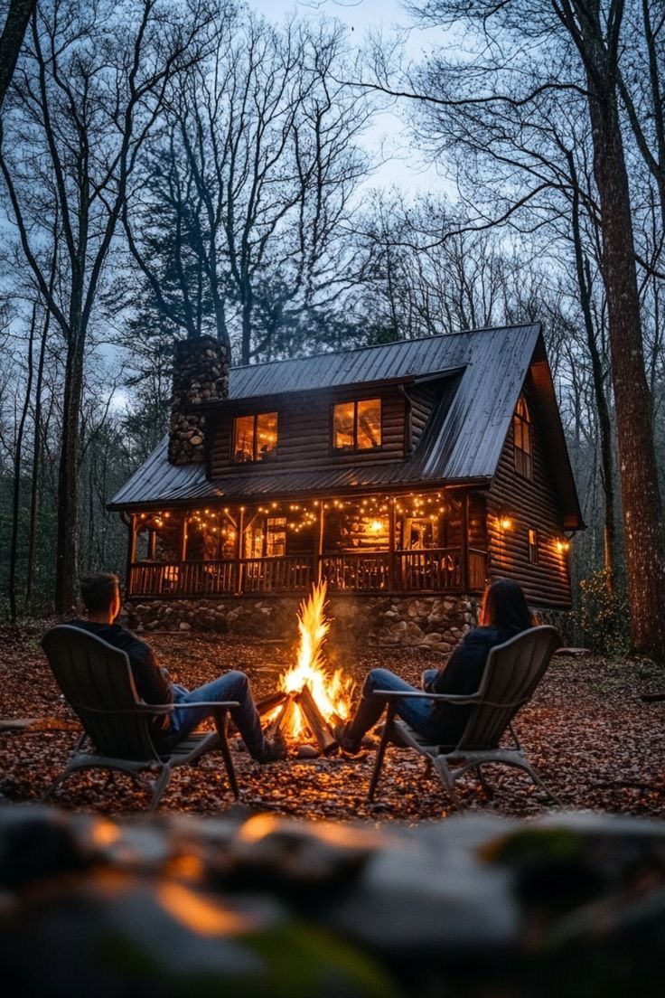 two people sitting in chairs next to a fire pit with a cabin in the background