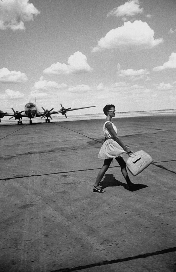 a woman walking across an airport tarmac with two planes in the background and one carrying a suitcase