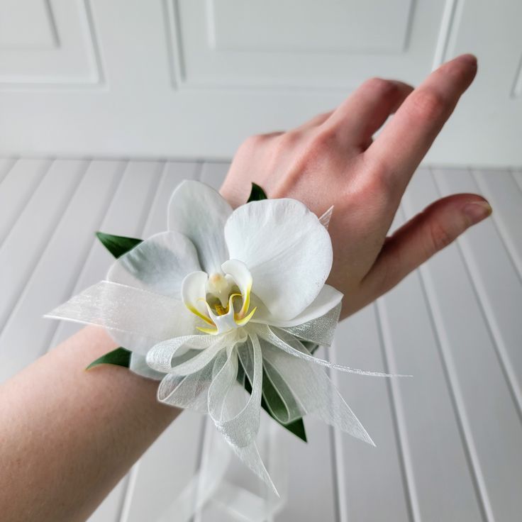 a person's hand holding a white flower with green leaves and ribbon on it