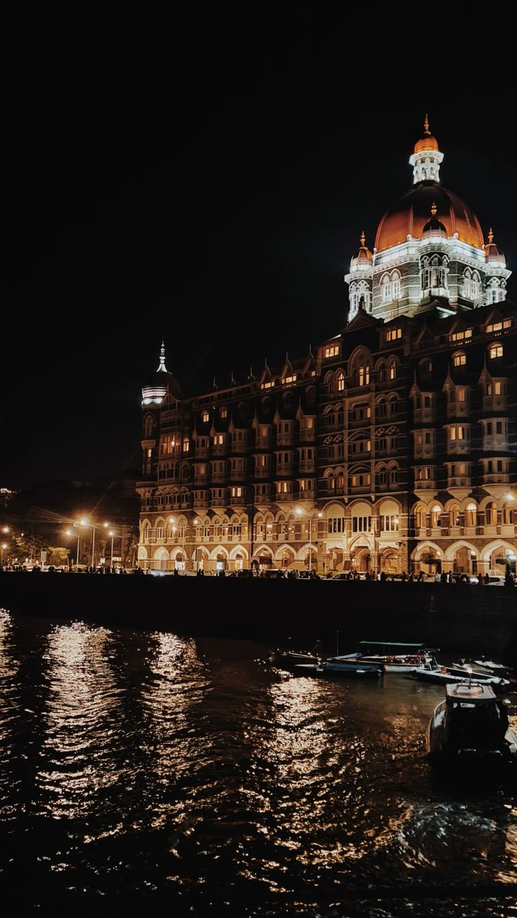 a large building lit up at night with boats in the foreground and lights reflecting on the water