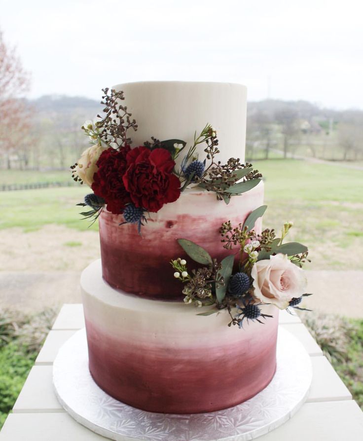 a three tiered cake with flowers and greenery on the top is sitting on a table outside