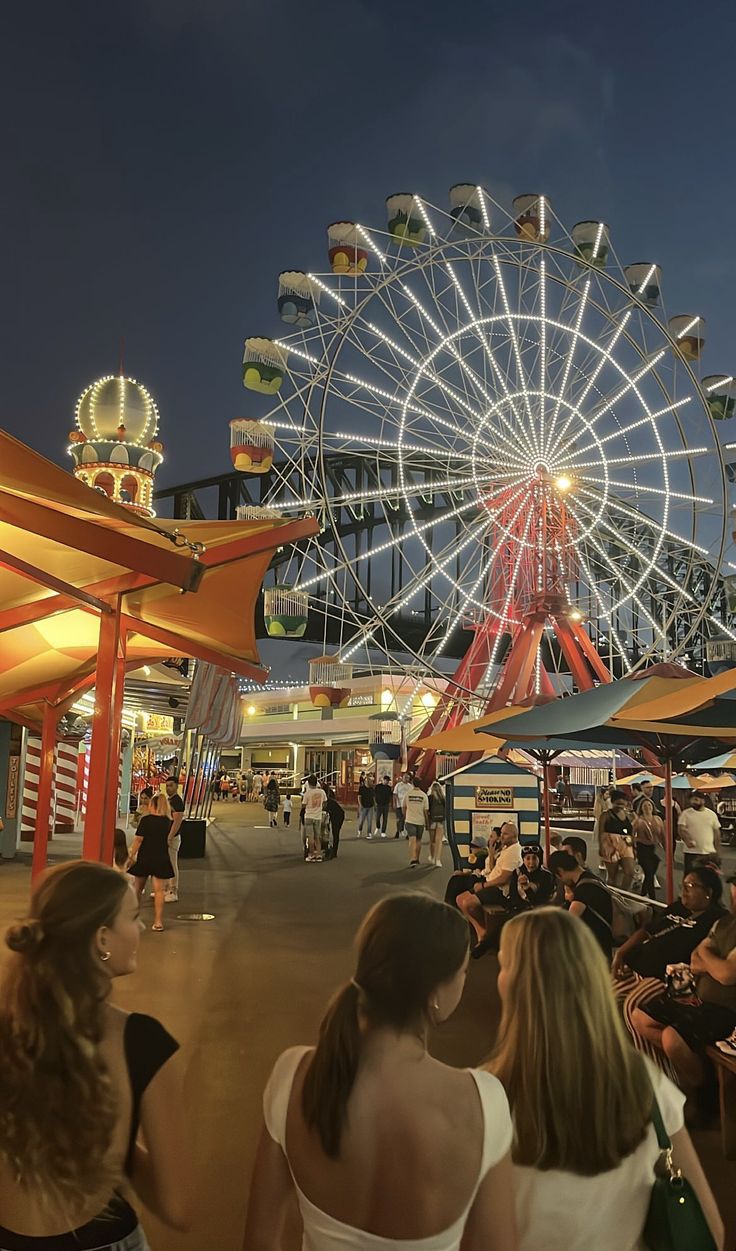 people are sitting and standing in front of a large ferris wheel at night with lights on