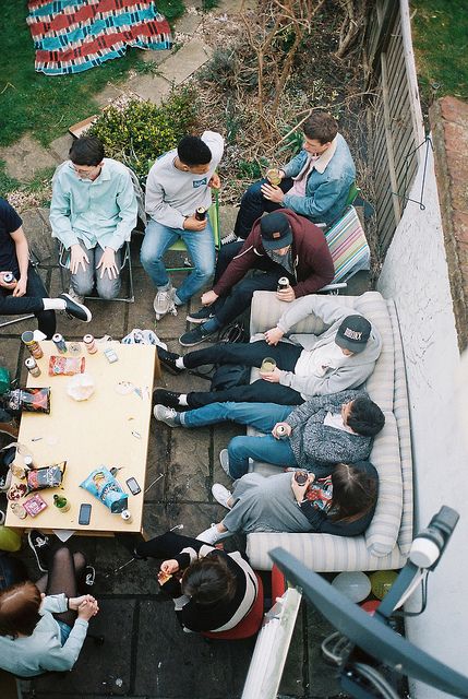 a group of people sitting around a wooden table eating food and drinking wine in the backyard