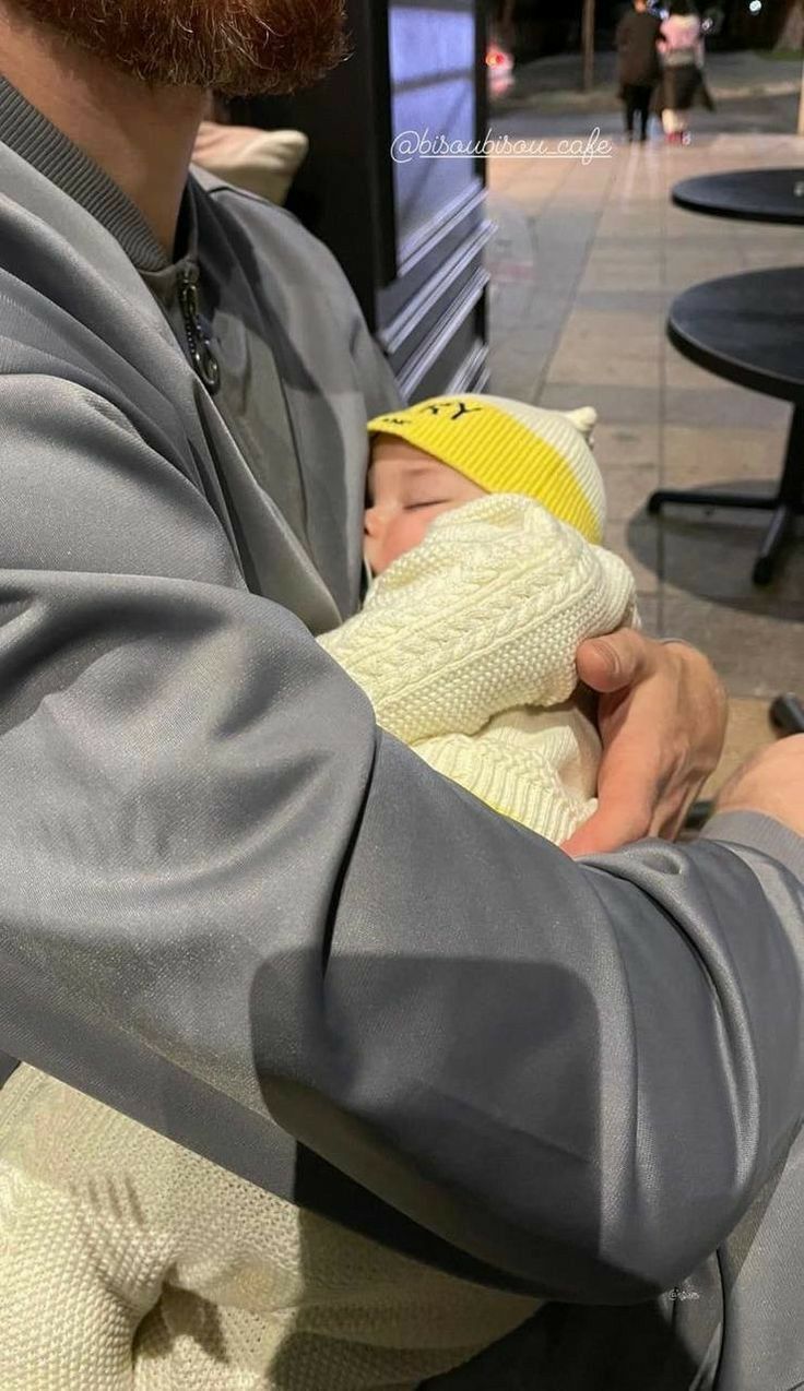 a man holding a baby wearing a yellow knitted hat and grey jacket with his arms wrapped around him