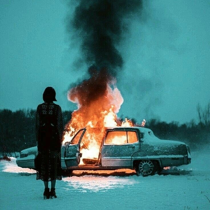 a woman standing next to a car on fire in the middle of snow covered ground