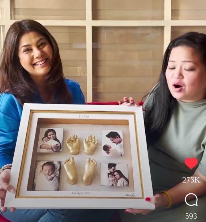 two women sitting on a couch holding up a framed photo with baby's handprints