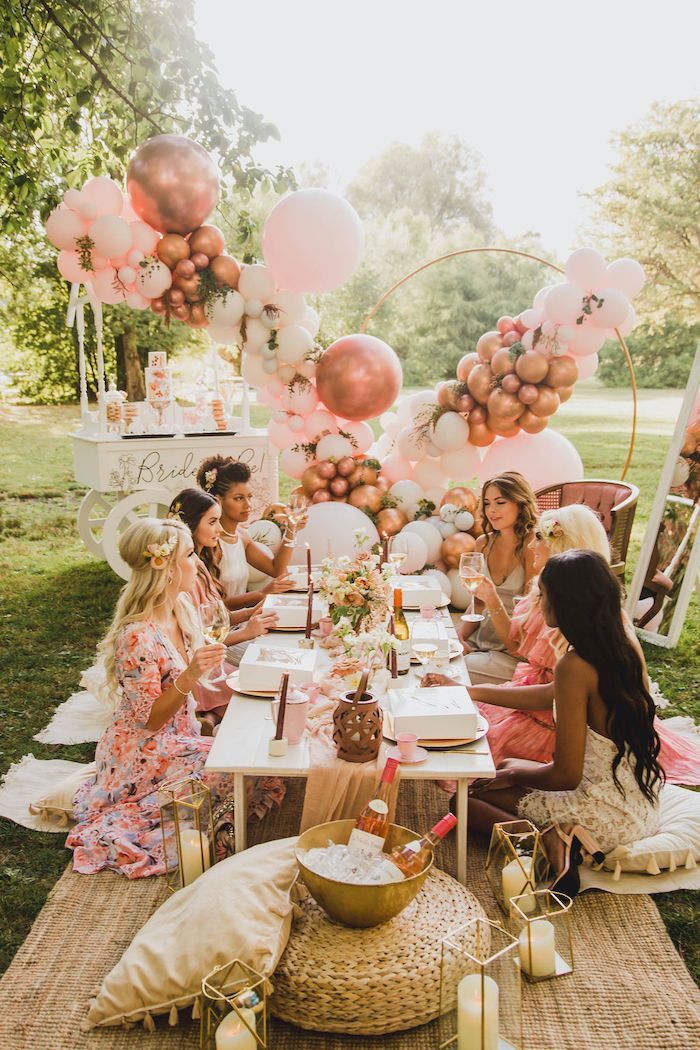 a group of women sitting at a table with balloons and plates on it in the grass