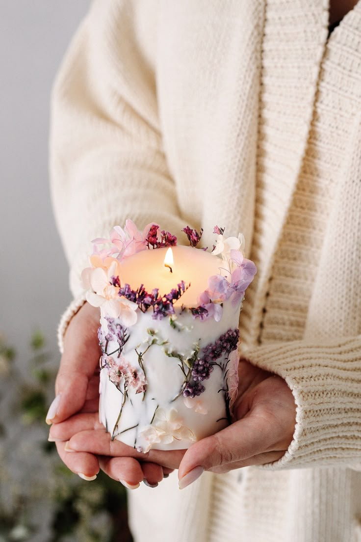 a woman holding a candle in her hands with flowers on it and a white sweater