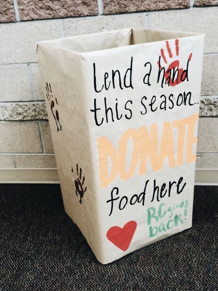 a cardboard box with writing on it sitting in front of a brick wall that says lend a hand this season, donation food here