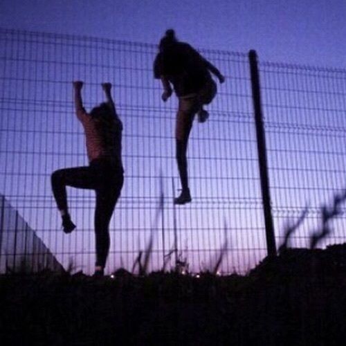 two people jumping in the air behind a fence at dusk or dawn with their arms up