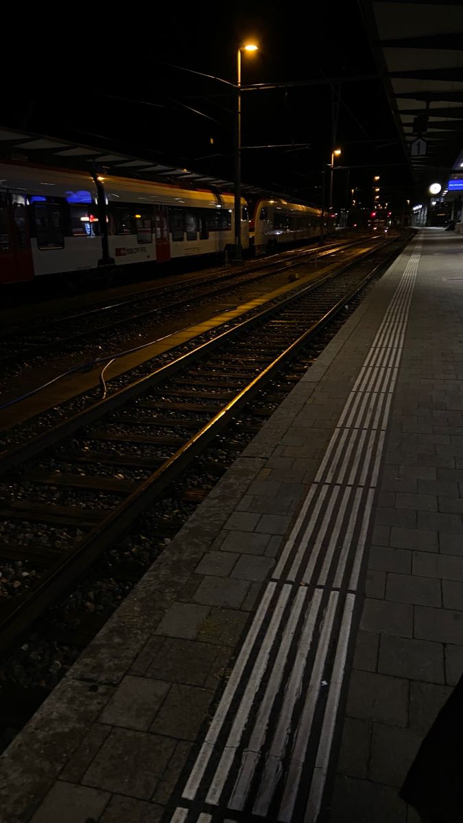 a train station at night with the lights on and tracks running parallel to each other