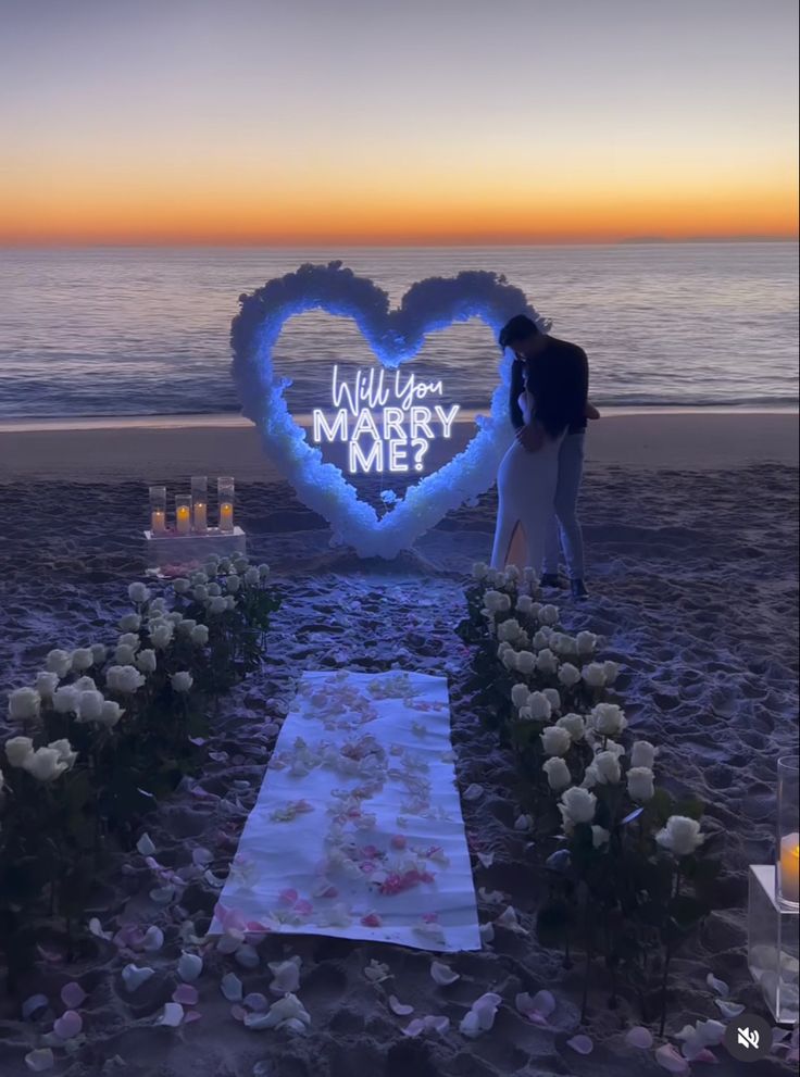 a man and woman standing in front of a heart - shaped sign on the beach