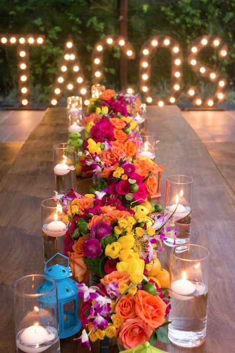 a long table with candles and colorful flowers on it, in front of some lights