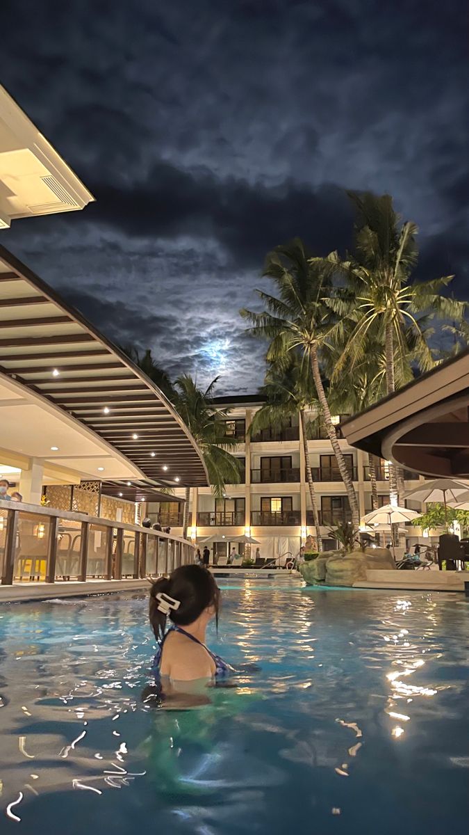 a woman sitting in the middle of a swimming pool at night with palm trees behind her