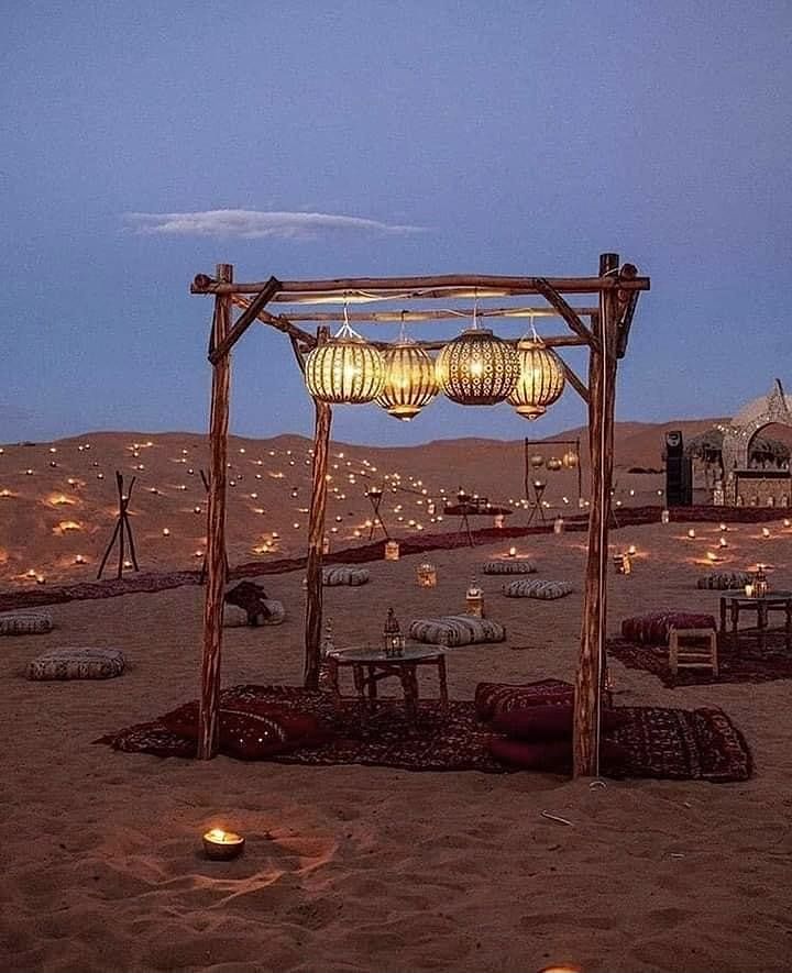 a desert setting with lanterns and tables on the sand, in front of an open fire pit