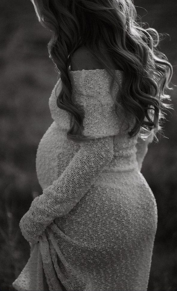 black and white photograph of a pregnant woman wearing a sweater in the grass with her hands on her hips