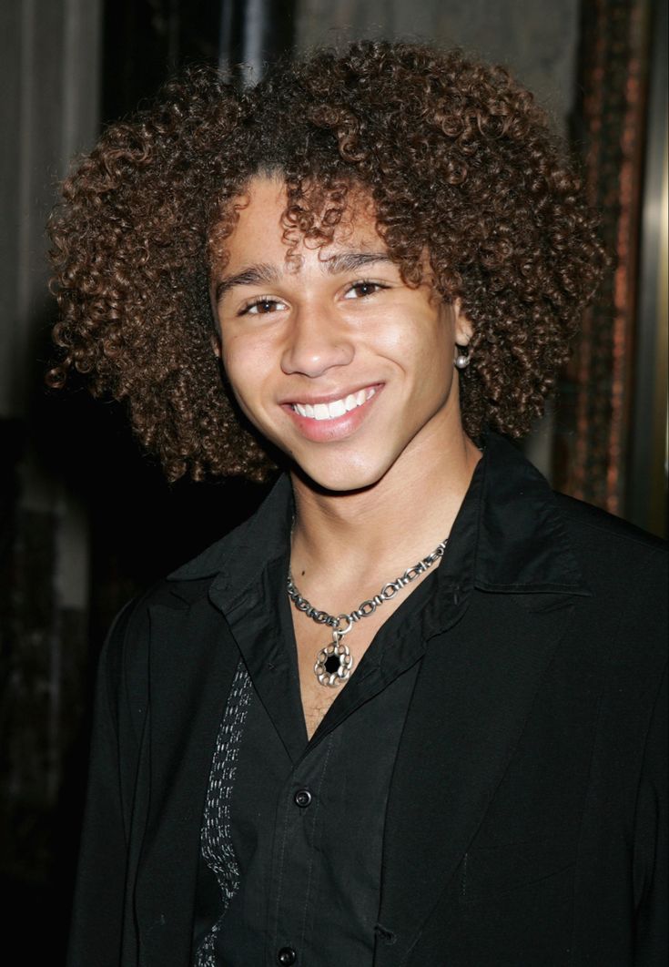 a young man with curly hair smiling at the camera, wearing a black shirt and silver necklace