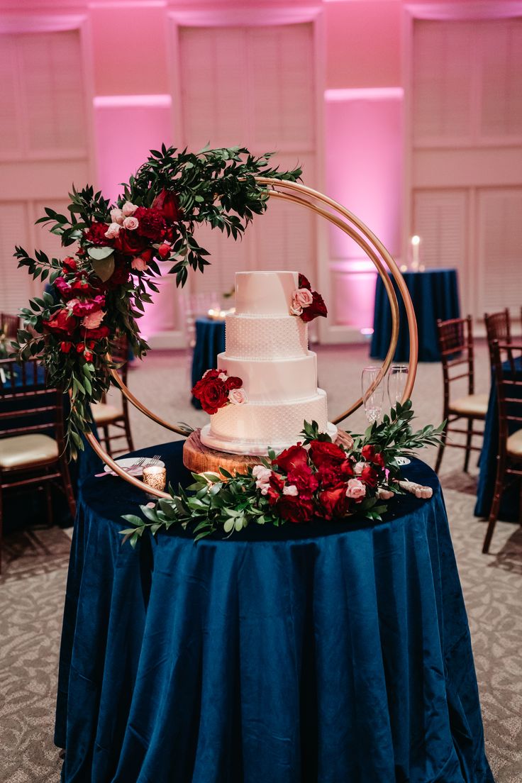 a white wedding cake with red flowers and greenery on top sits on a blue table cloth