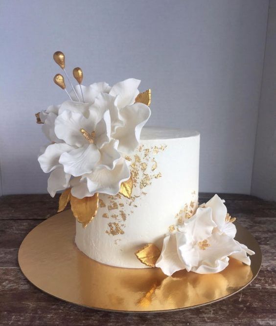 a white and gold wedding cake with flowers on the top is sitting on a wooden table