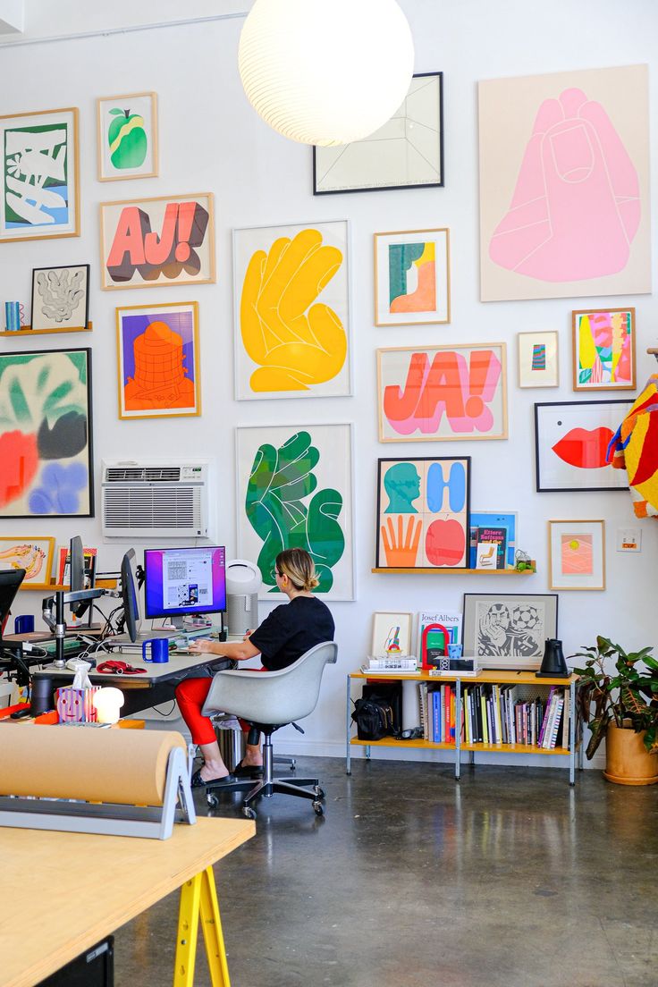 a woman sitting at a desk in front of a wall full of pictures and artwork