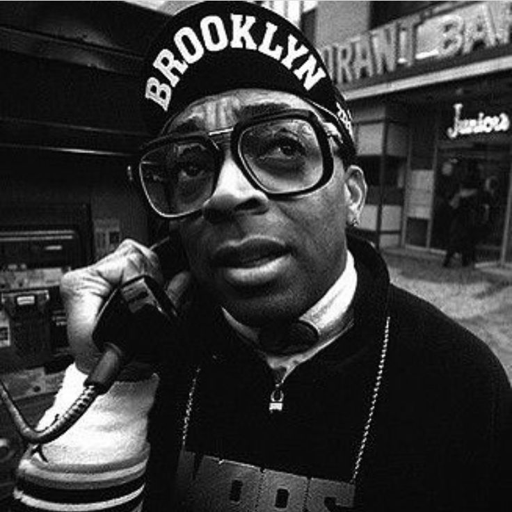 a man wearing glasses and a hat talking on a cell phone while standing in front of a telephone booth