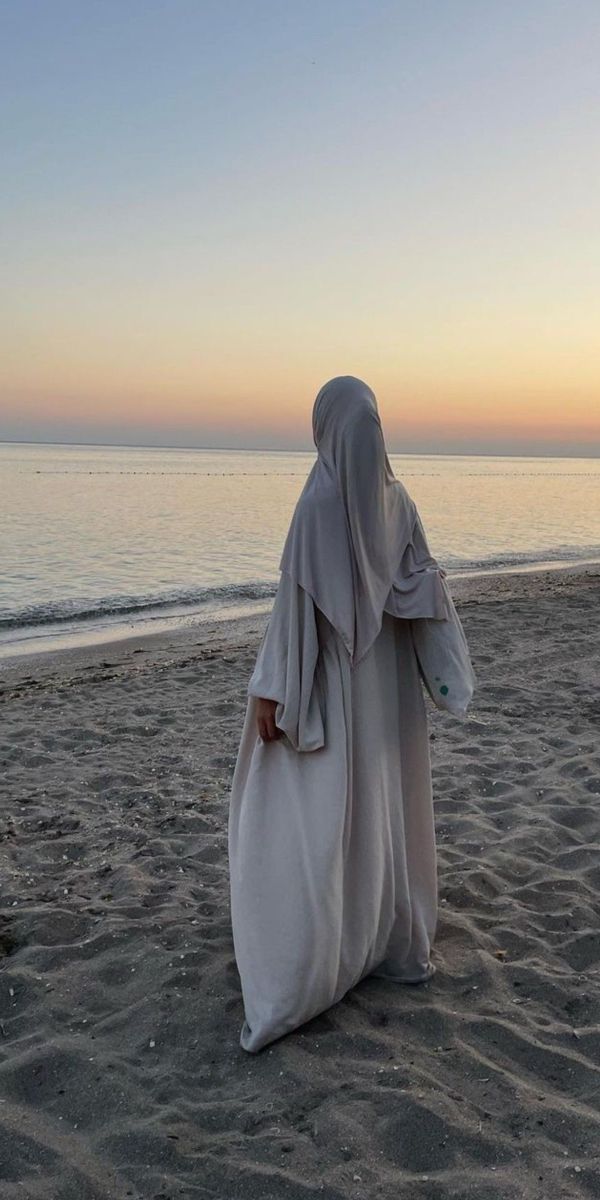 a woman in a white robe walking on the beach at sunset with her back to the camera