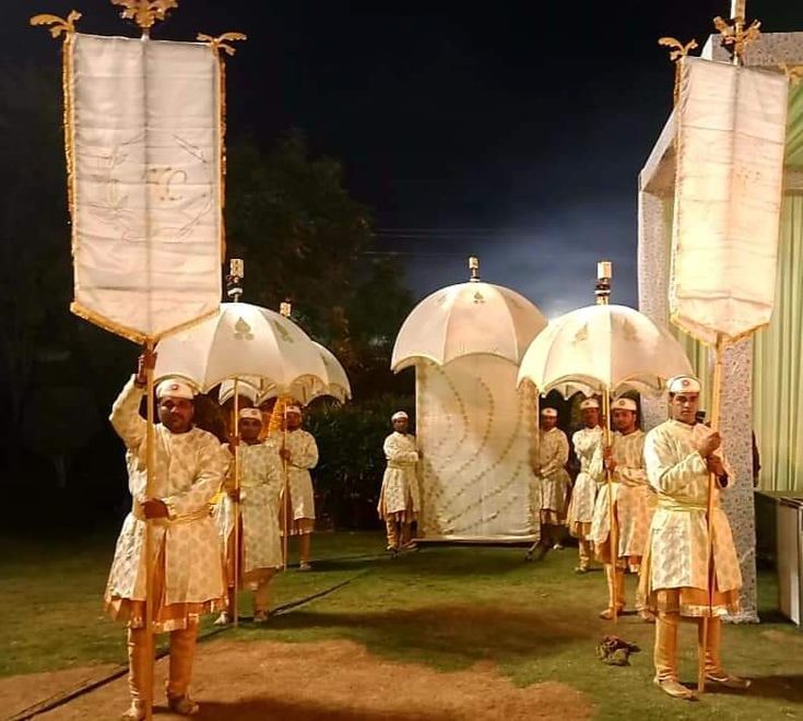 several men dressed in white and gold holding umbrellas while standing on grass with other people behind them