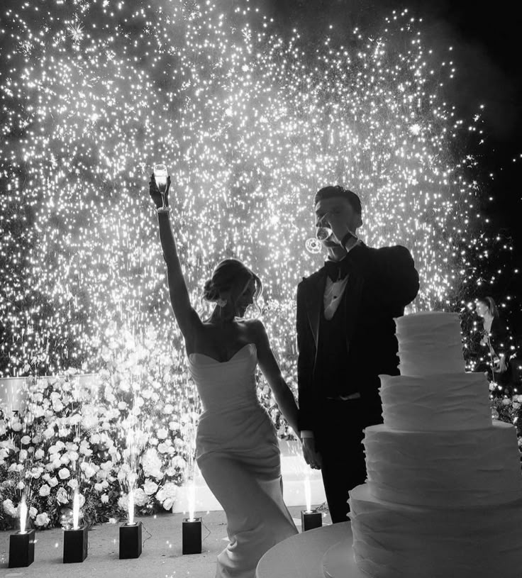 a bride and groom standing next to a wedding cake with fireworks in the background at night