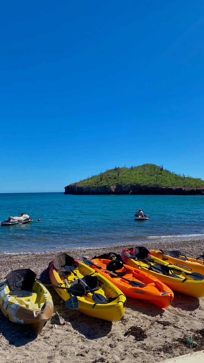 several kayaks lined up on the beach with people in boats behind them and an island in the distance