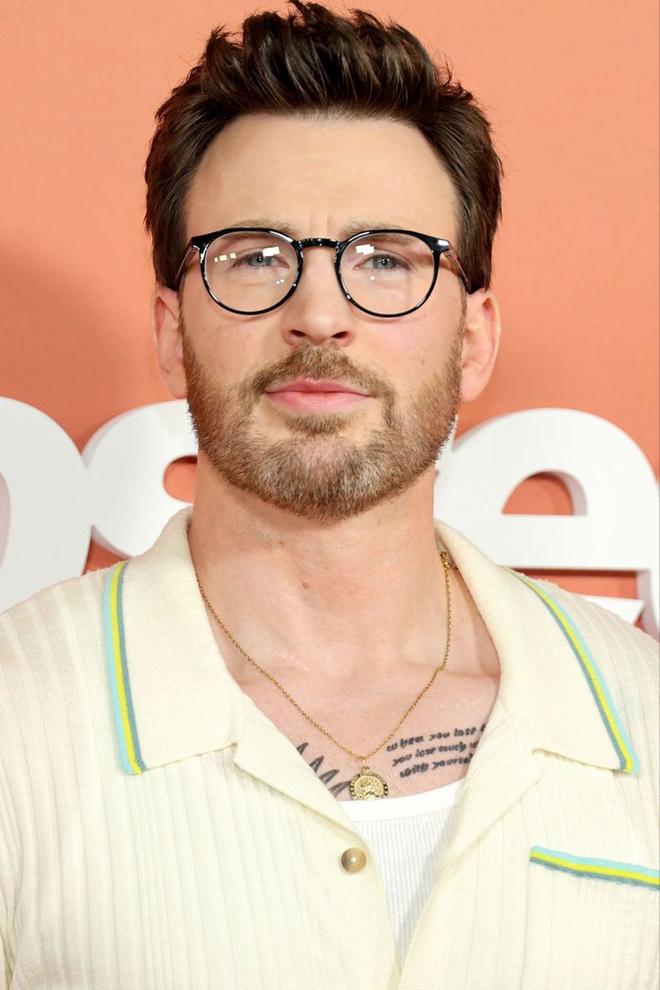 a man with glasses and a beard in front of a pink wall wearing a white shirt