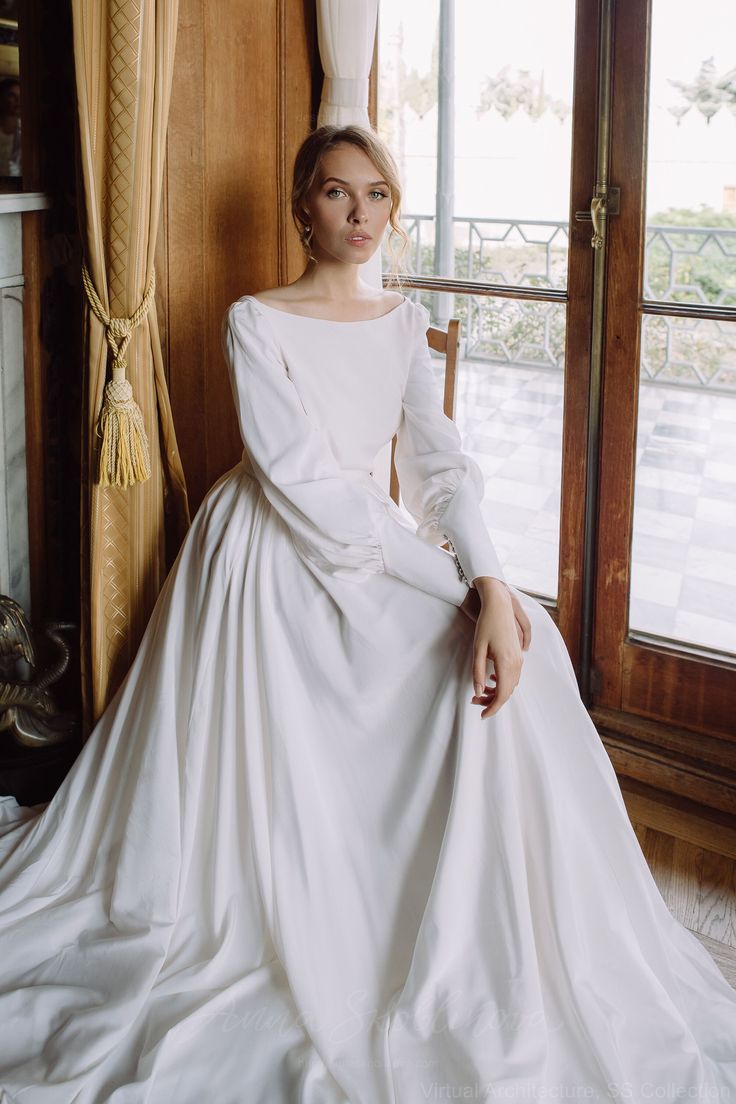 a woman in a white wedding dress sitting on a chair by a window with her hands on her hips