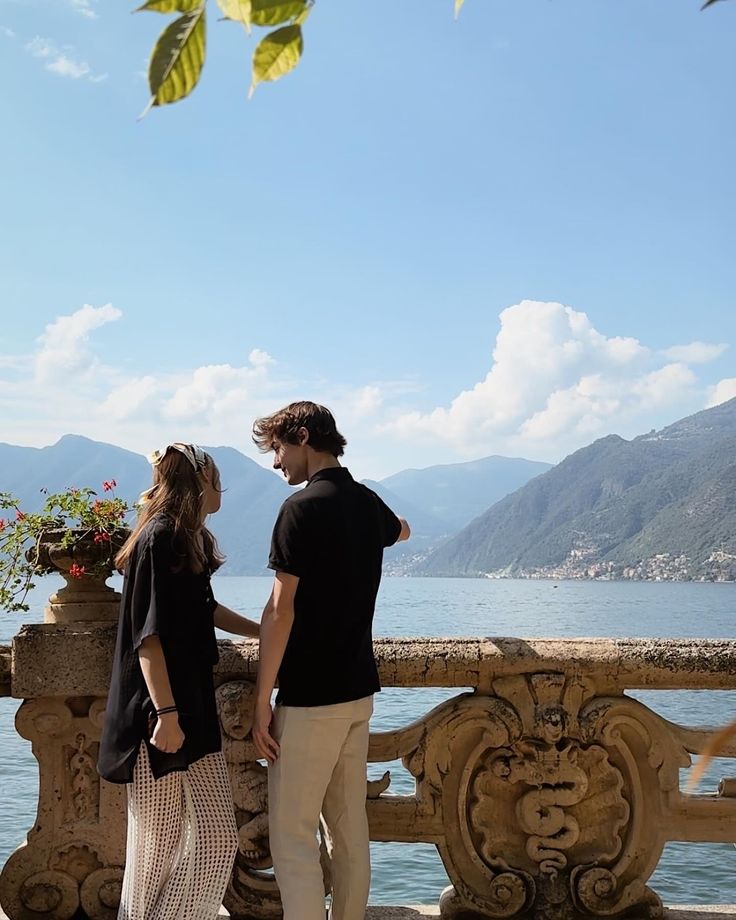 a man and woman standing next to each other on a balcony near the water with mountains in the background