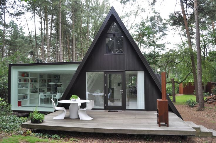 a small black house in the woods with tables and chairs on the front porch, surrounded by trees