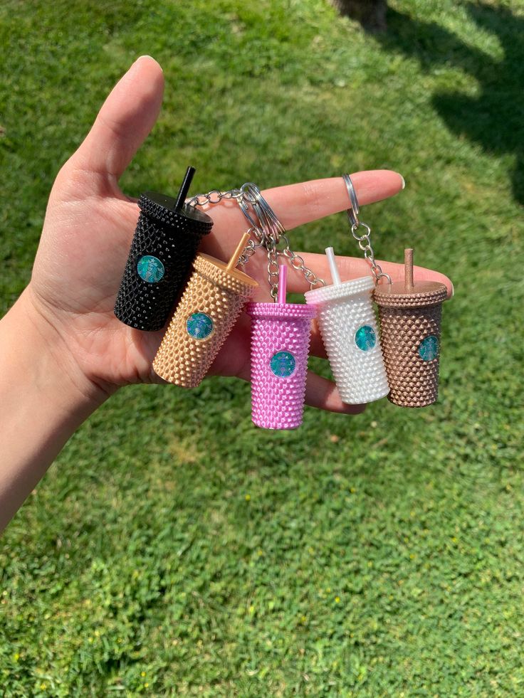 a person holding four different colored cups in their hand on the grass with one being held up by two fingers