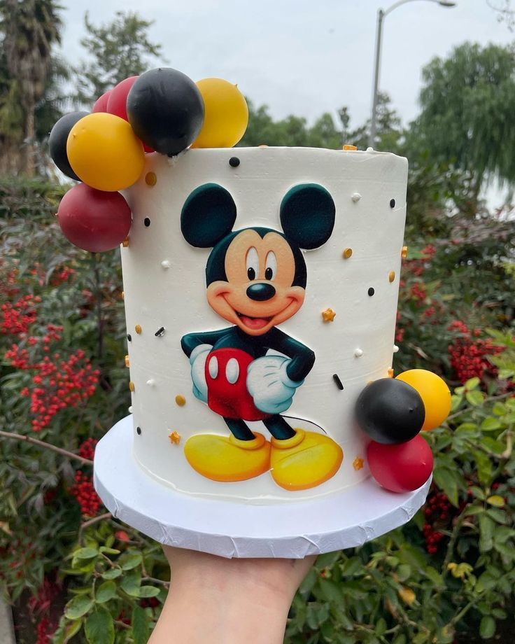 a hand holding a mickey mouse cake with balloons on it's top and bottom