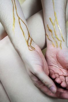 two hands with gold leaf tattoos on their arms and fingers, both holding each other