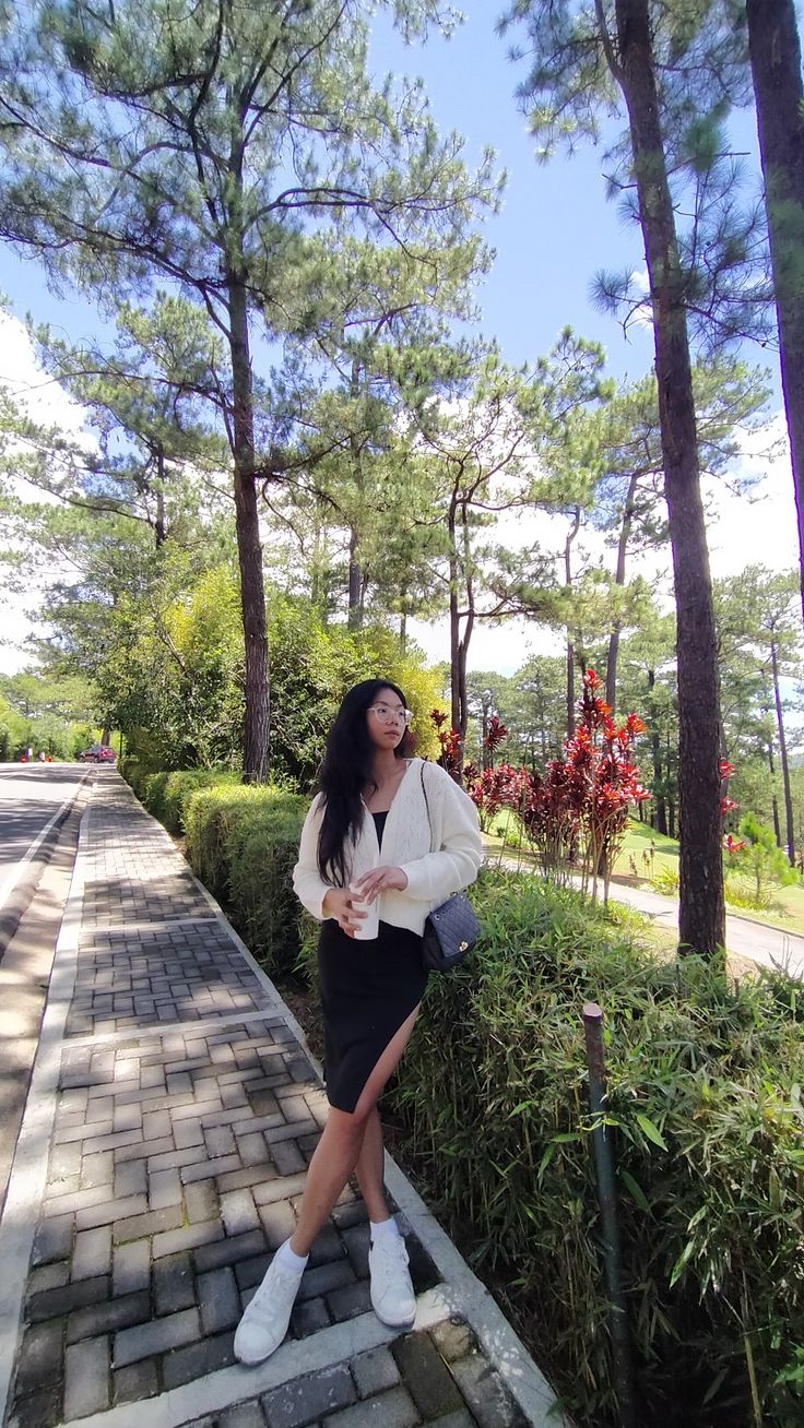 a woman in white shirt and black skirt standing on brick walkway next to bushes with trees