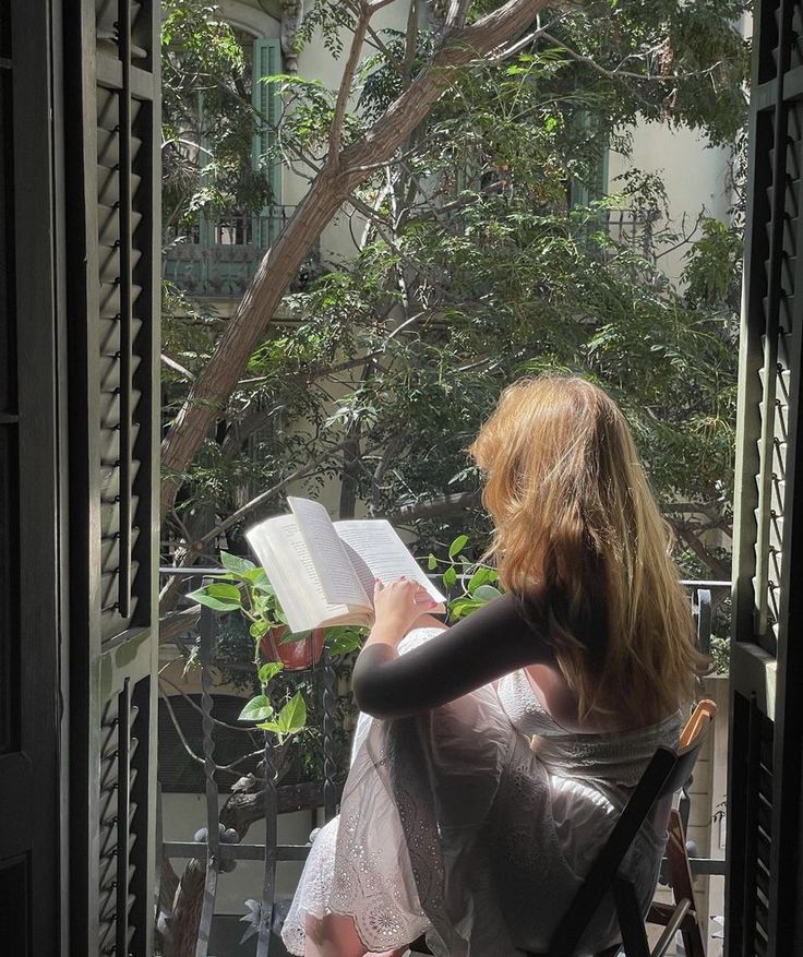 a woman sitting in a chair reading a book on a balcony with trees and bushes behind her