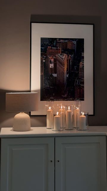 lit candles sit on top of a white cabinet in front of a framed cityscape