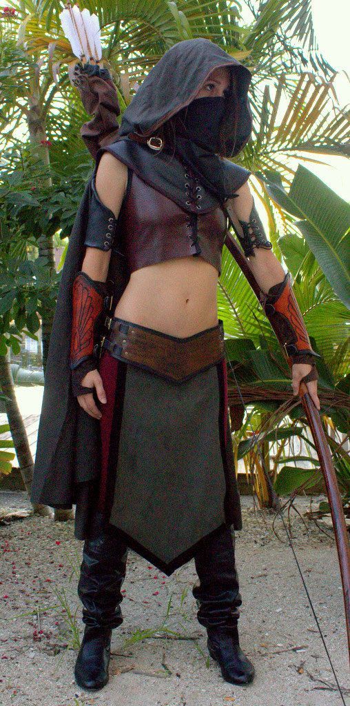 a woman dressed in costume holding a bow and arrow while standing on the ground with palm trees behind her