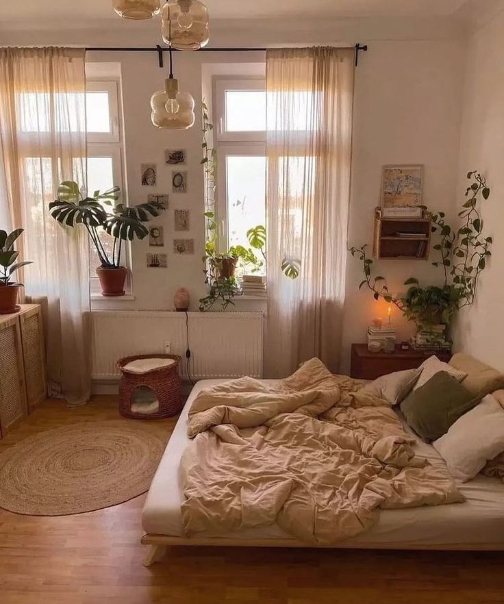 an unmade bed sitting in a bedroom next to two windows with plants on them