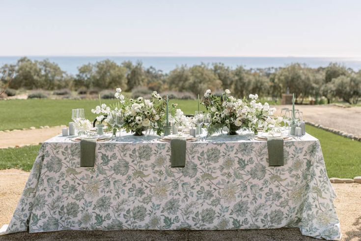 the table is set with white flowers and greenery on it for an outdoor wedding reception