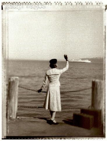 an old photo of a woman standing on a dock waving at the ocean with a boat in the distance