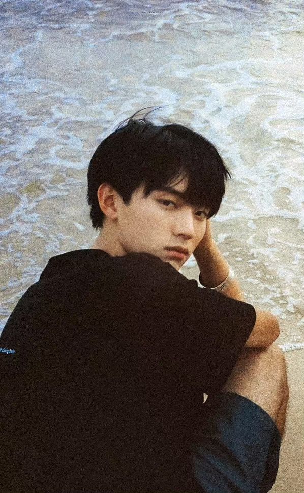 a young man sitting on top of a sandy beach next to the ocean with his hand under his chin