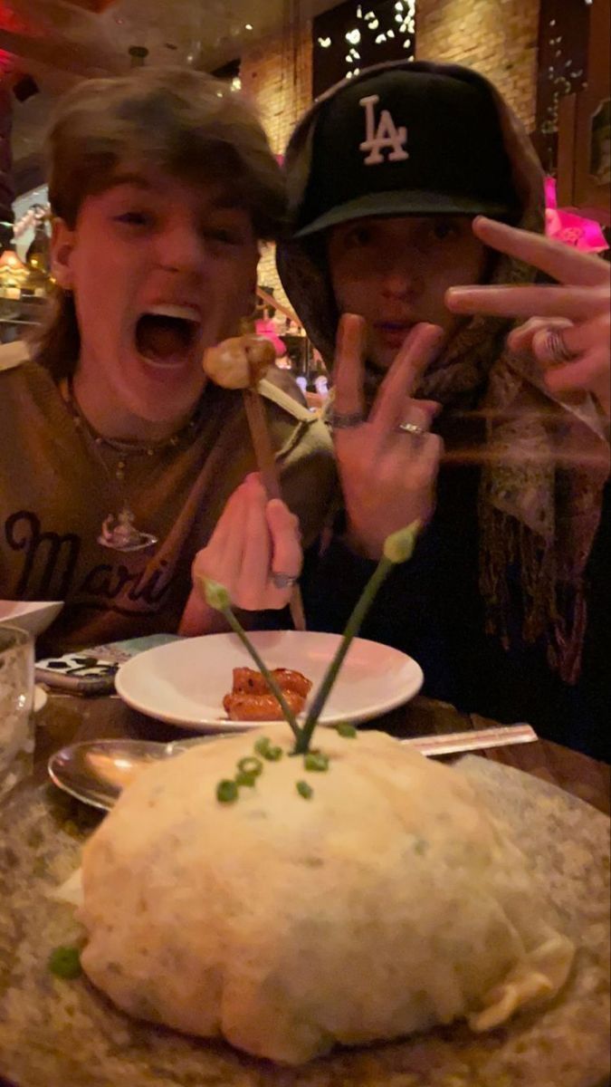 two people sitting at a table with food in front of them and one person making the peace sign