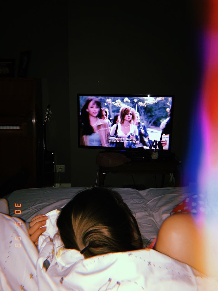 a woman laying in bed watching tv with her head on the pillow and hands behind her back