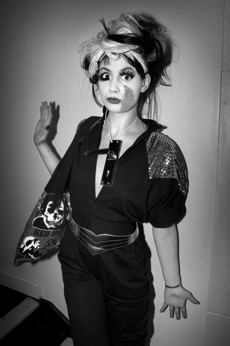 a woman with makeup on her face and hands in the air while standing next to a wall