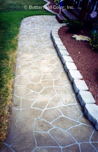 a stone walkway in front of a house with grass and flowers on the other side