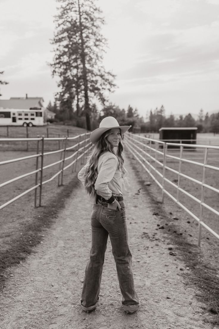 a woman standing in front of a fence wearing a cowboy hat and jeans with her hands on her hips